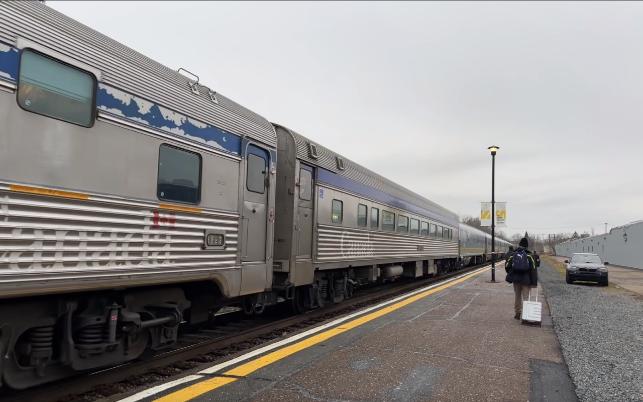 A man with a black jacket pulling a silver carry-on suitcase walks along a paved platform with his back to the camera, apparently heading to board the rather decrepit grey passenger train parked on his left, under a pale grey sky.
