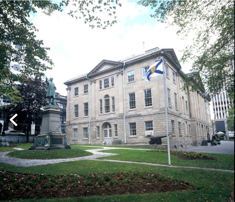 A historic-looking, three-storey stone building with tall, elegant windows,framed by a few tree branches, and fronted by green lawn and garden, with a winding path leading towards the building. Credit: Communications Nova Scotia