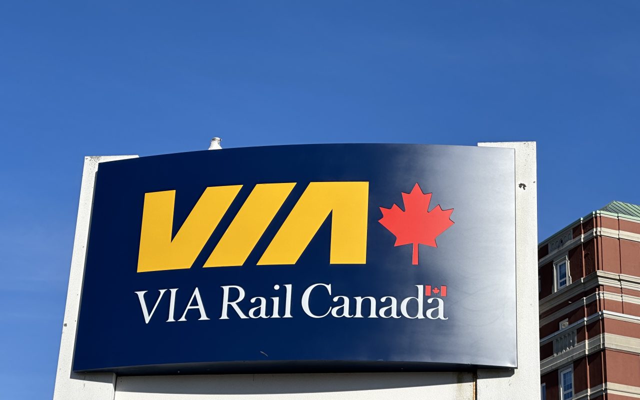 Large sign with dark blue background, and VIA in yellow letters, then a red maple leaf, and underneath the words "VIA Rail Canada" in white lettering. Behind the sign, a red brick building and a very blue sky. A white seagull sits atop the sign.