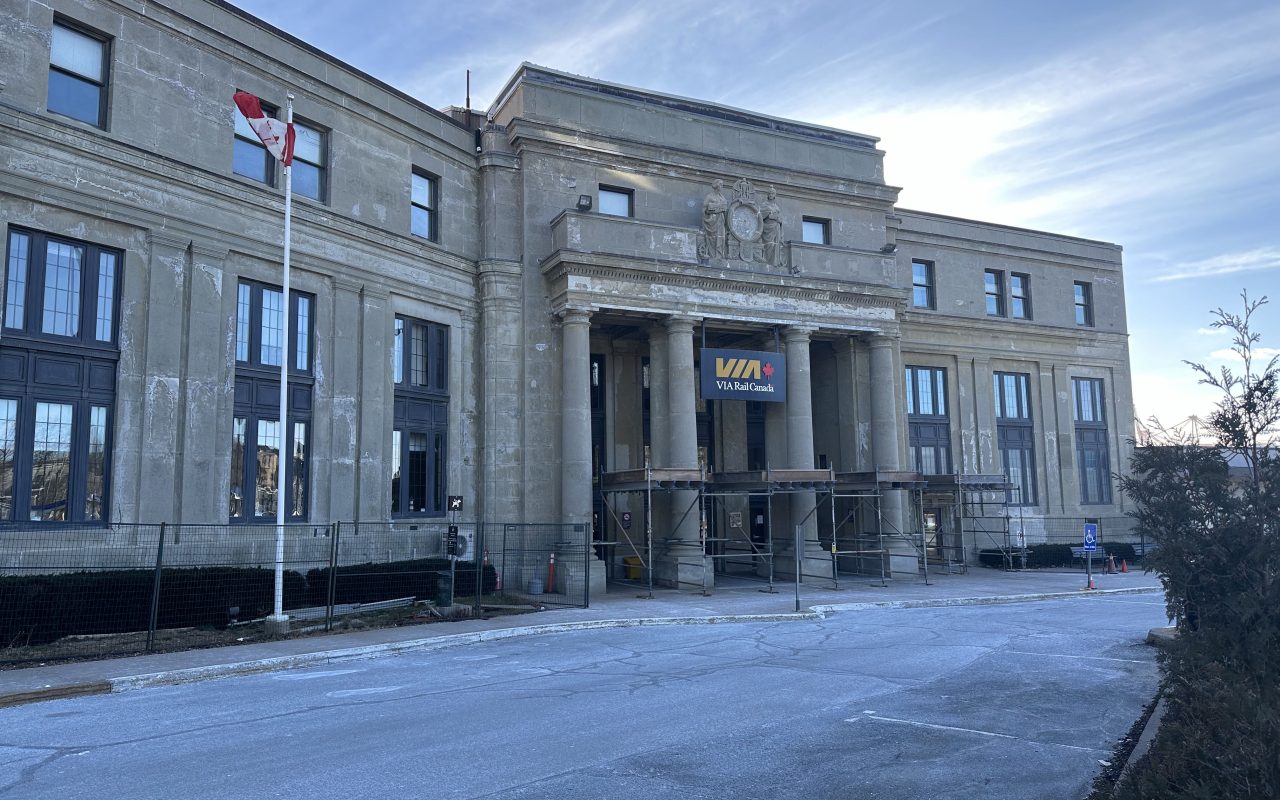 Concrete facade of an imposing building, looking a little worse for wear, with a Canadian flag flying to the left of the columns and entrance to the building, over which there is a blue sign with the yellow logo of VIA Rail and its red maple leaf.